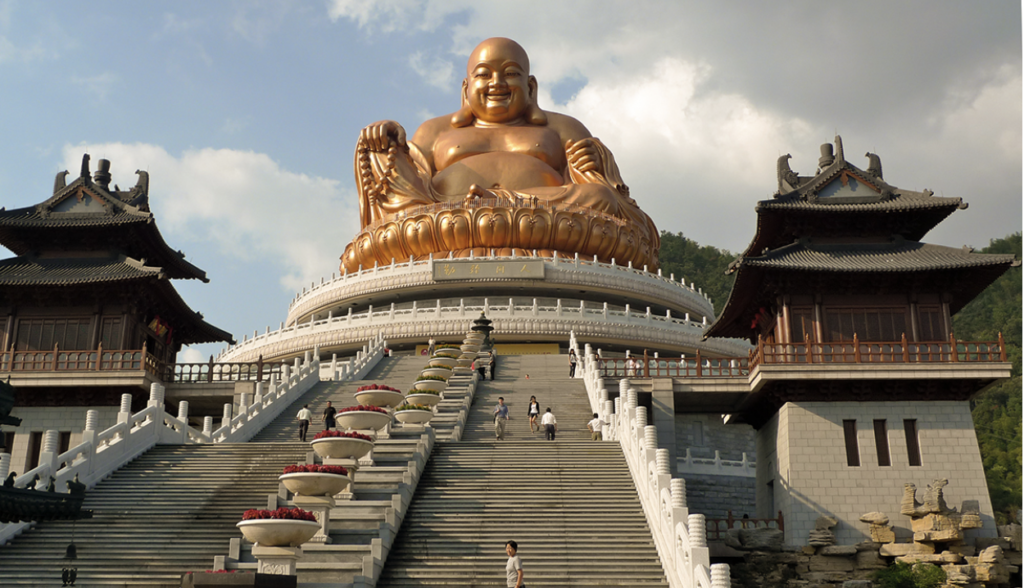 A Buddhsit temple with two towers on both sides with many stairs with a white fence on both sides and red flowers in white pots in the middle, leading to the temple which is in a two white ring structure with a gold Buddha on top looking upon all that enter the temple.