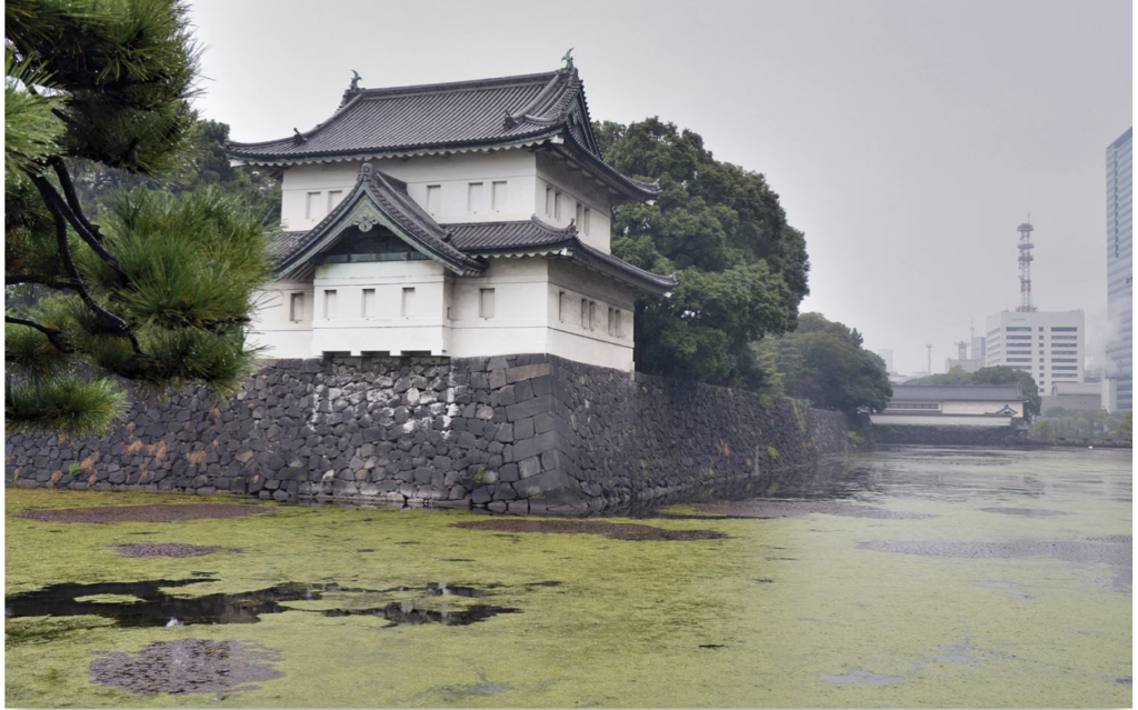 A white building with oriental type roofing that seems to overlook a river. It could be considered a watchtower. There are many windows looking out into the river and it is sitting on a stone foundation that seems to be weathered. It also backs a forest that has a bridge connecting to modern buildings to the right of it.